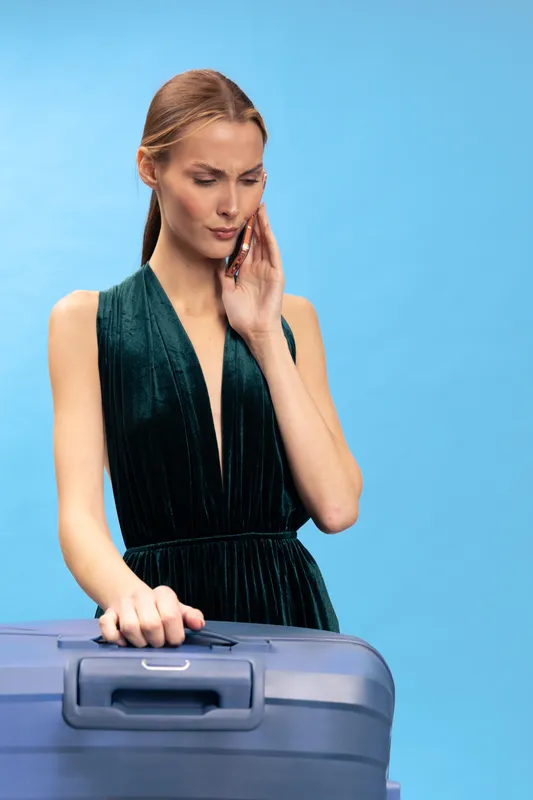 Concerned young woman in elegant dress holds a suitcase and talks on phone with serious expression. Blue background. Concept for travel problems, communication and stress.