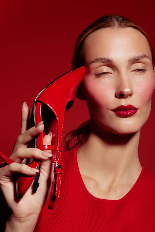 Woman with red lipstick and red dress holds red high heel shoe near face with closed eyes. Concept of fashion, beauty, style, and elegance on red background.