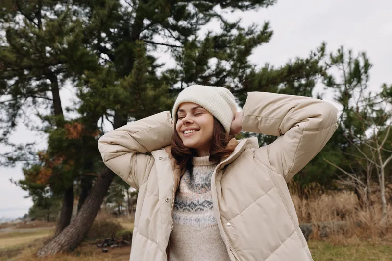 Outdoor woman in a cozy winter jacket and sweater, smiling with arms behind head in a windy landscape of trees and grass, relaxed lifestyle scene.
