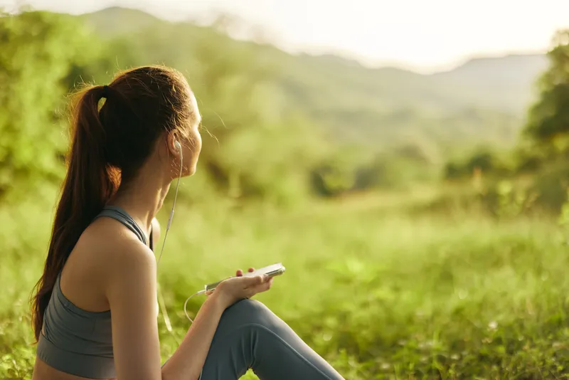Young woman enjoying nature sitting on grass, wearing athletic wear, looking at the mountains, conveying calmness and relaxation in a lush green environment
