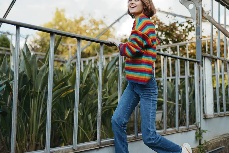 Teenage girl in a rainbow sweater and jeans smiling while walking along a metal railing in an urban botanical setting, casual outfit, short brown hair, happy lifestyle portrait.