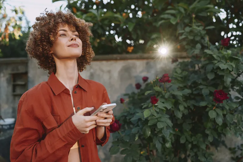 Cheerful woman outdoors, looking up, soft sun flare, curly hair, casual orange shirt, holding smartphone, lush garden setting, warm natural colors, relaxed lifestyle moment