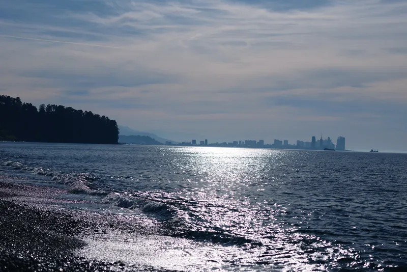 Water, lake, sea and skyline with sun reflection across calm shoreline and distant city silhouette on the horizon over sparkling waves, peaceful coastal mood.