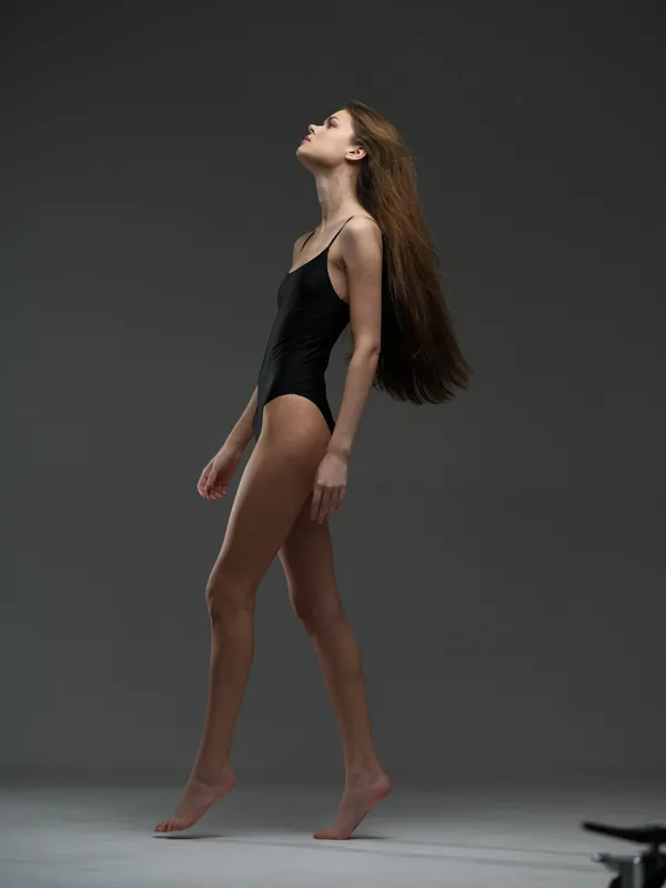 slender woman in black bodysuit walking gracefully with closed eyes and long hair on grey background