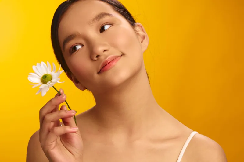 Cheerful asian woman holds a delicate daisy against a vivid yellow backdrop, soft studio lighting, a gentle smile, and a calm, inviting mood that conveys warmth and charm.