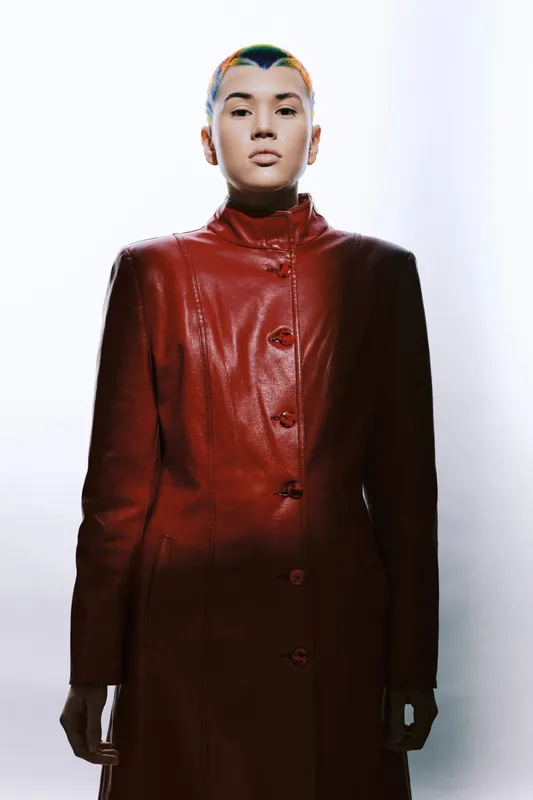 Woman with short colorful hair wearing a red leather coat stands confidently against a bright white background, showcasing modern fashion and minimalistic style.