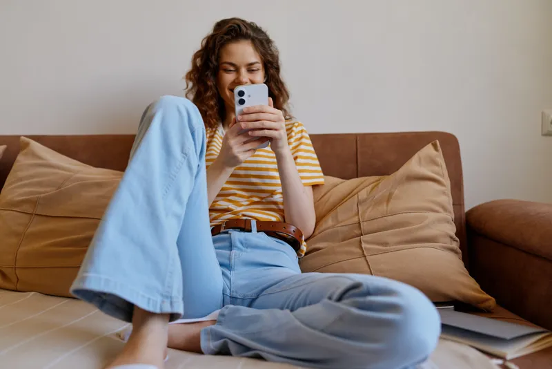 Casual woman indoors, relaxed on a couch, scrolling on a smartphone, striped tee and jeans, natural light, cozy living room, soft cushions, warm tones, comfortable home atmosphere, candid lifestyle
