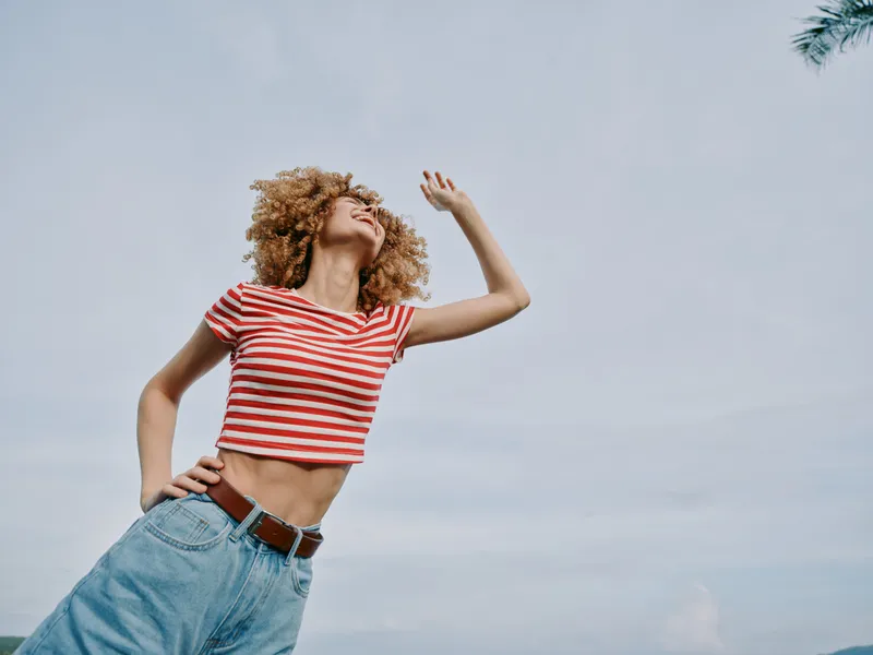Young woman with curly hair wearing a red and white striped crop top and denim jeans stands outdoors under a bright sky, captured in vibrant movement and joy.