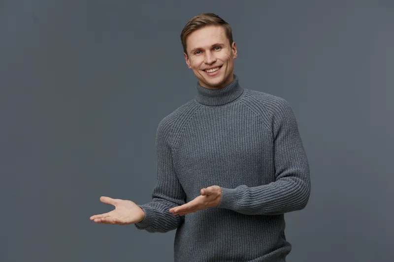 smiling caucasian man in gray turtleneck sweater showing presenting gesture isolated on gray background mental health and gadgets concept