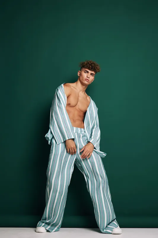 Young man standing wearing blue and white striped fashion clothing shirt open and trousers with casual white shoes on green background. Model with curly hair posing full length portrait.