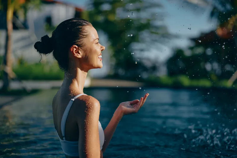 Young woman with dark hair in a bun enjoys splashing water while standing in an outdoor pool on a sunny day, expressing happiness and relaxation in a lifestyle setting.