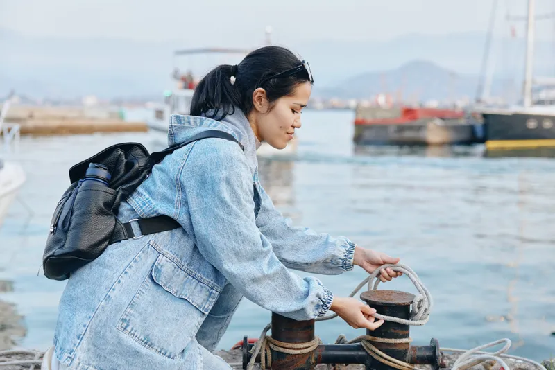 Female traveler in a denim jacket by the harbor adjusts the mooring rope, with a backpack and boats in the distance, capturing a calm outdoor maritime moment