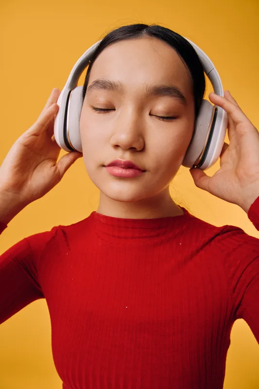 Young woman with closed eyes wearing headphones listening to music, enjoying sound in casual red top against orange background for relaxation and entertainment.