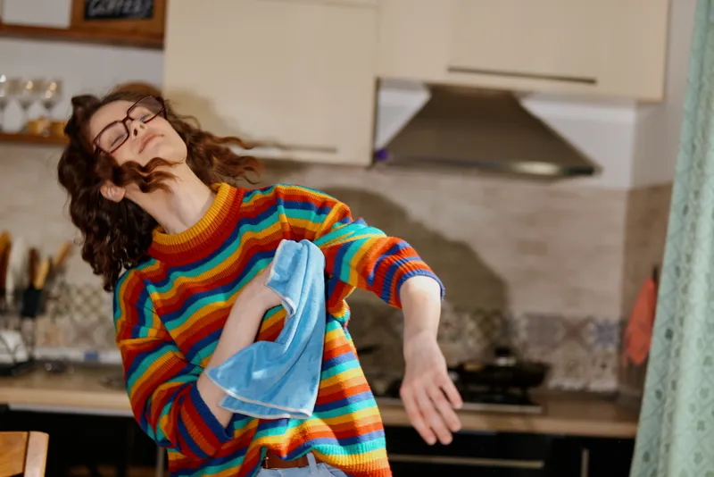 A woman in a colorful striped sweater wipes a cloth in a bright kitchen, capturing a spontaneous home moment, casual daily routine, and the warmth of everyday life.