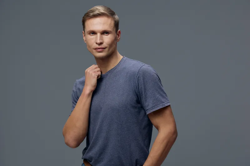 Confident young man with short light brown hair wearing casual blue t-shirt looking directly at camera in studio portrait. Male fashion and style concept