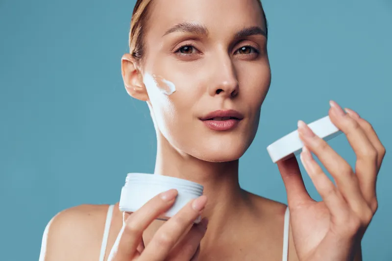 Young woman applies face cream from jar on cheek with gentle expression, skincare routine concept, clear skin, isolated on blue background, beauty and self care, closeup portrait.