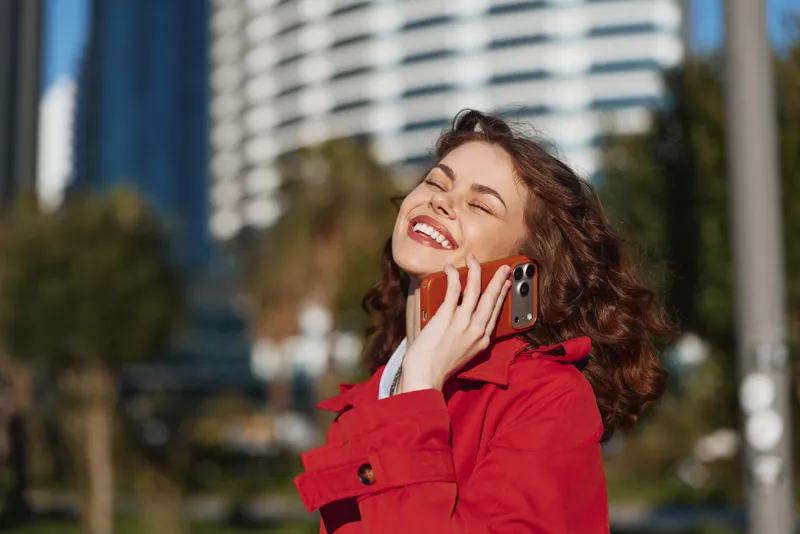 Young woman with curly hair smiles joyfully while talking on a smartphone outdoors in a red coat on a sunny day in an urban park setting.