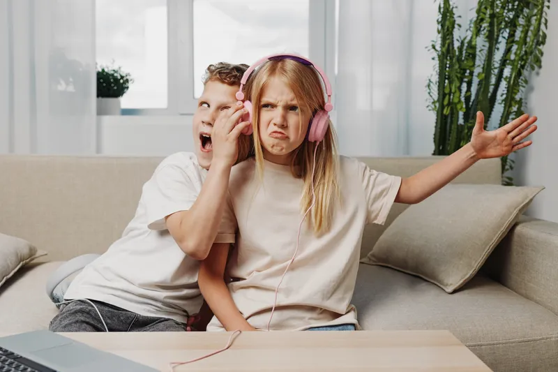 Annoyed girl with headphones refuses to share music while boy yawns loudly sitting on couch in bright living room interior.