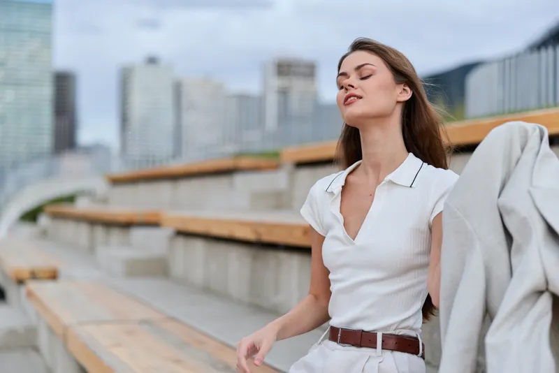 Young woman with closed eyes and relaxed expression sitting outdoors in casual white outfit with city skyline in soft background, natural light, spring style