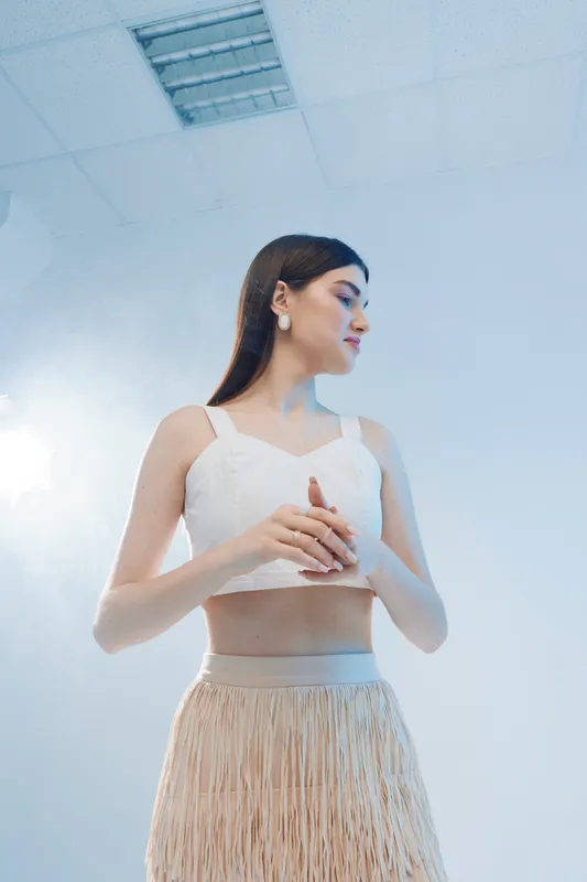 Young woman in white top and beige skirt poses gracefully in a bright studio. Her long hair and elegant earrings highlight beauty and fashion concepts with soft lighting.