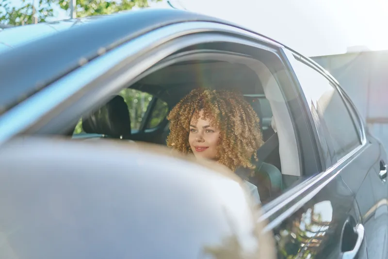 Young woman with curly hair sitting in a modern car, smiling softly with natural sunlight and green trees outside, casual and relaxed mood.