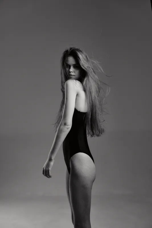 Black and white portrait of a young woman with long hair wearing a black bodysuit, posing with a serious expression in a studio setting.