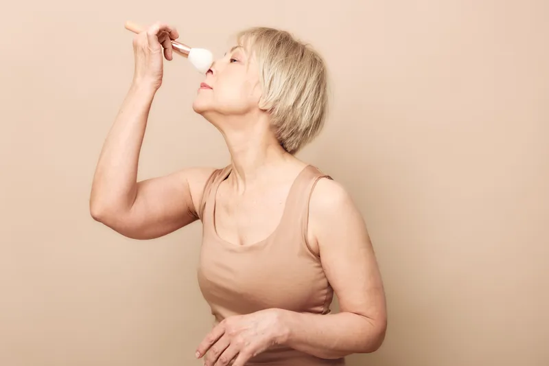 Mature woman with short blonde hair applies makeup powder on face with brush, wearing beige sleeveless top against neutral background. Concept of beauty, self-care, and aging gracefully.