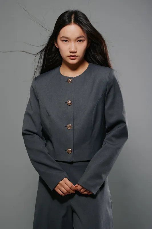 Young woman with long black hair wearing gray jacket and matching pants standing against gray background. Serious expression and poised posture in studio portrait fashion style.