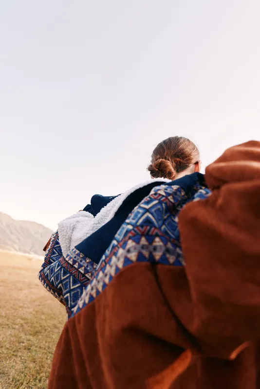 Person with patterned blanket and jacket outdoors in mountains, back view showing cozy layered textile and travel vibe across a scenic field landscape at golden light.
