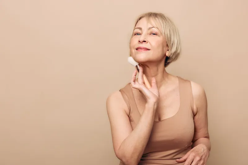 Mature woman with short blonde hair applies soft makeup brush on face, smiling gently in beige top against neutral background. Concept of beauty, skincare, and self-confidence.