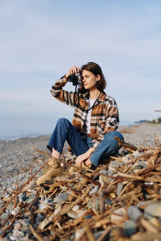Photographer seated on a rocky beach with a camera raised, casual plaid shirt and jeans, capturing coastal landscape and distant waves in an outdoor creative shot.