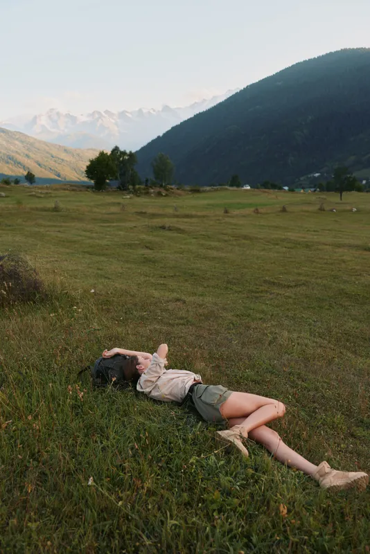 Young woman with dark curly hair lies relaxed on green grass in a wide open meadow surrounded by mountains under a clear sky.