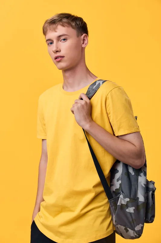Man wearing a yellow shirt with a camo backpack against a bright yellow studio backdrop, casual youth style, ready for school or travel, fashion forward and ready for daily urban exploration