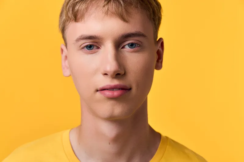Close up of a young man with a calm expression against a bright yellow backdrop, clean skin, short hair, and casual style in a minimal studio setting.