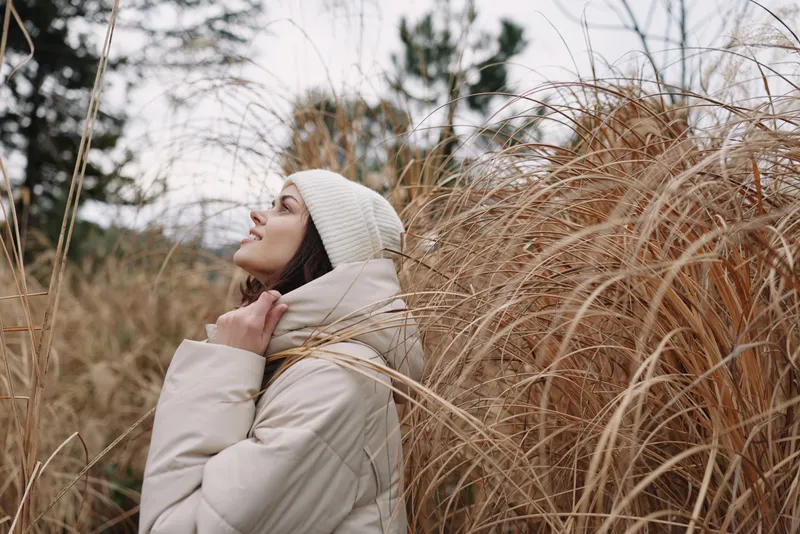 Winter portrait of a woman in a beige coat, scarf and beanie, standing among tall dry grasses in a quiet field, delivering a peaceful, cozy and reflective mood.