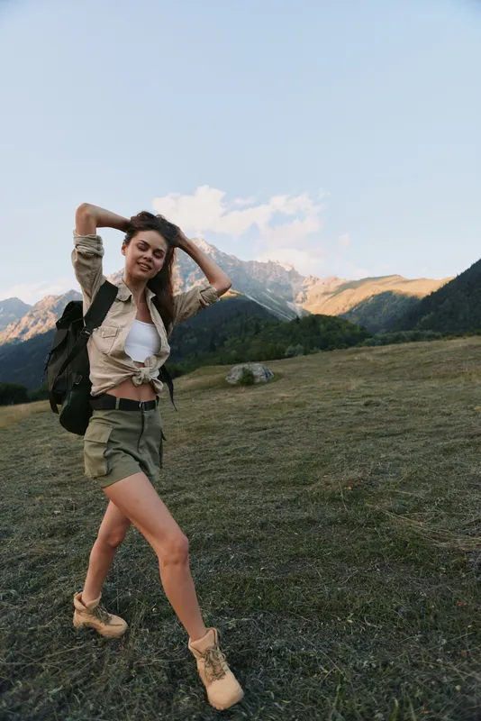 Young woman hiking through a meadow with mountains in the background, backpack on her shoulder, enjoying a carefree outdoor adventure as she explores the rugged trail.