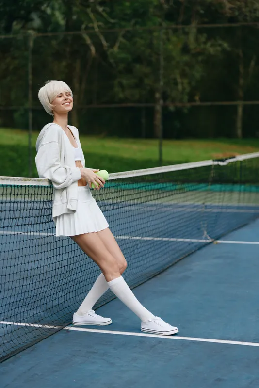 Young female tennis player in white sportswear smiling on a court surrounded by greenery, showcasing a healthy and active lifestyle perfect for sports related content