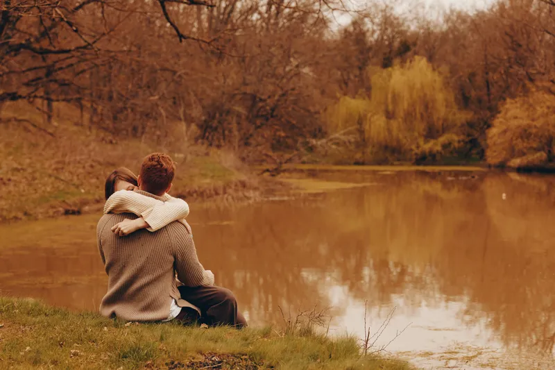 Couple embrace on grassy bank beside a calm lake, autumn colors surrounding them, conveying tenderness and companionship in a peaceful outdoor moment. Isolated setting, warm light, soft breeze,