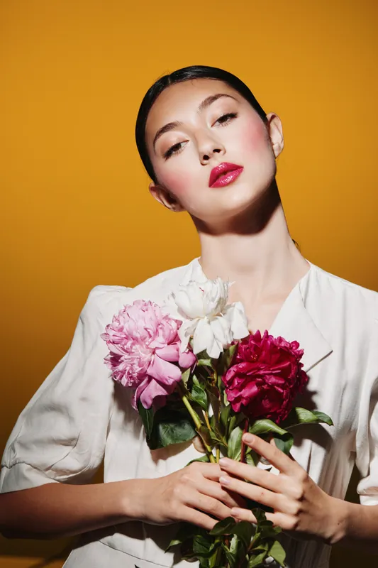 Fashion model with bright red lipstick poses with peonies against orange background wearing white blazer. Beauty creative concept for trend bridal or elegant style.