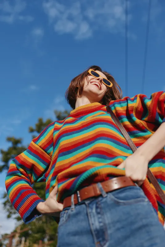 woman smiling striped sweater yellow sunglasses jeans standing outdoors sunny, young adult in her 20s wearing casual outfit and leather belt, hands on hips, joyful travel lifestyle portrait under