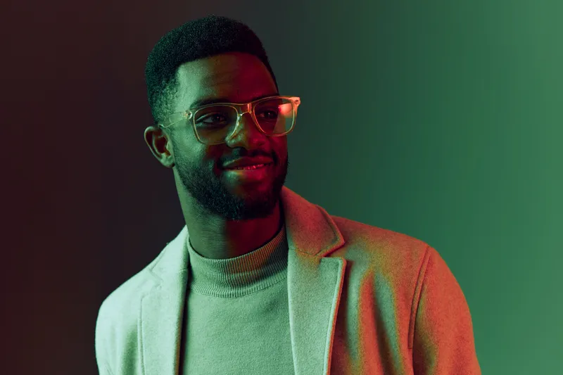 Smiling young Black man in stylish attire with glasses against a gradient green background showcasing modern fashion and confidence