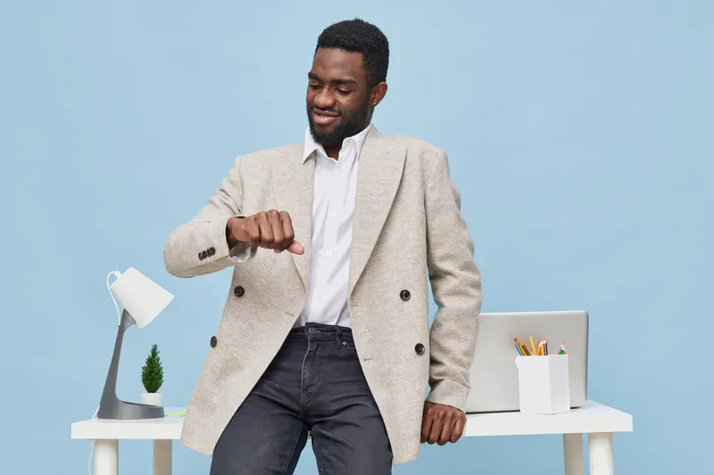 Confident man in beige blazer and white shirt standing in modern office with desk and laptop, looking at watch, smiling, relaxed, professional appearance, indoor setting, business casual style, bright