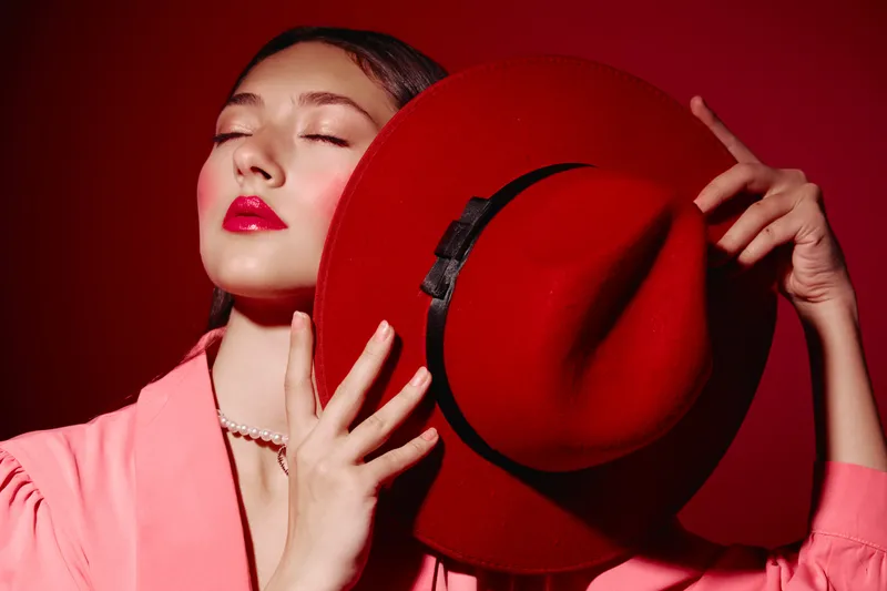 Fashionable young woman with glossy red lips posing with matching red hat and pearl necklace on red background. Artistic beauty portrait emphasizing style and luxury.