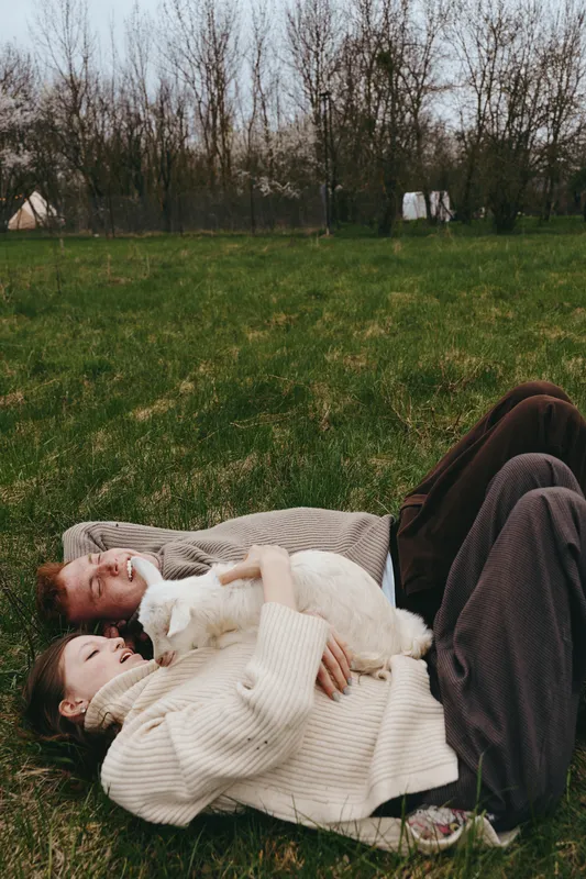 A couple lies on the grass in a relaxed outdoor scene, embracing a fluffy cat and sharing a warm smile, savoring simple lifestyle moments in a tranquil field, natural light and gentle breeze around