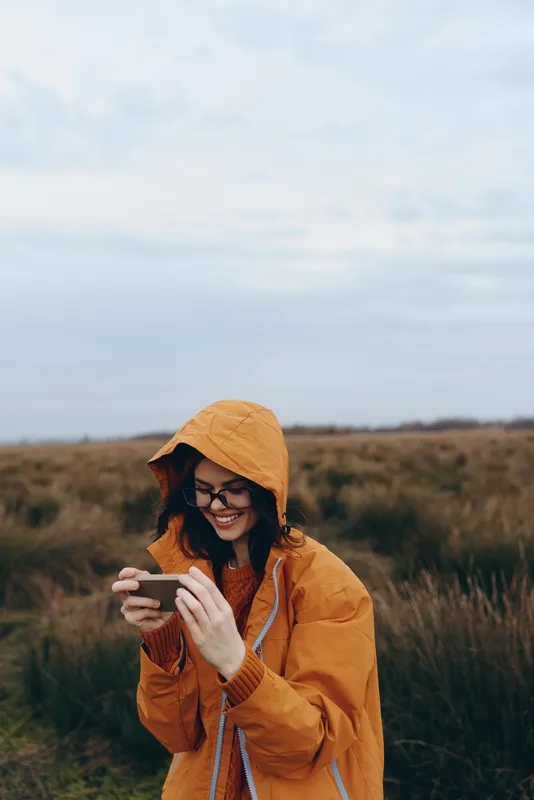 Smiling woman in orange raincoat with hood and glasses in an open field, holding smartphone and looking down, outdoor lifestyle in nature under daylight, calm everyday moment
