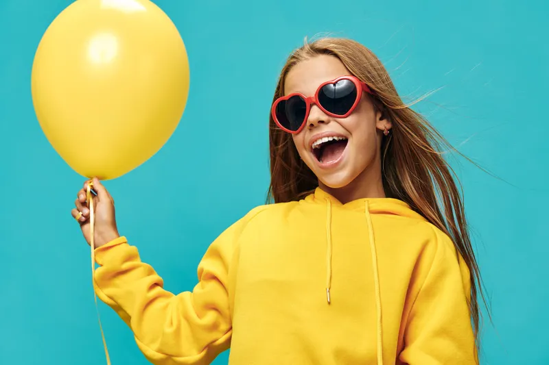 Girl in yellow hoodie with heart shaped sunglasses holding yellow balloon smiling on blue background, emotion of joy and celebration with casual style clothing.