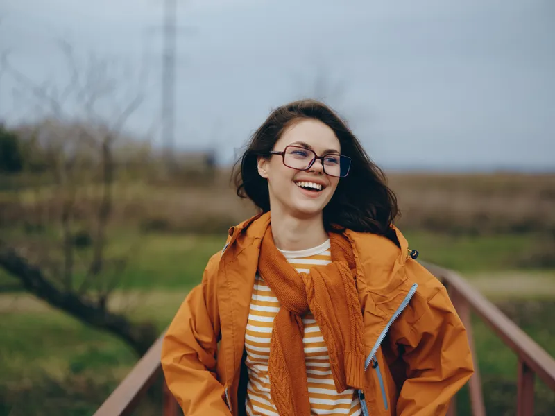 Smiling woman in orange coat and scarf, wearing glasses, outdoors on a countryside path with wooden railing, cloudy sky and wind in hair, lifestyle portrait in fresh air, during spring day