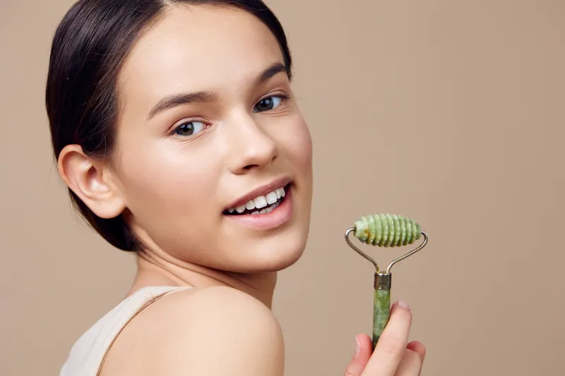Young woman smiling while holding a jade roller, showcasing healthy skincare routine with minimalistic background and natural makeup