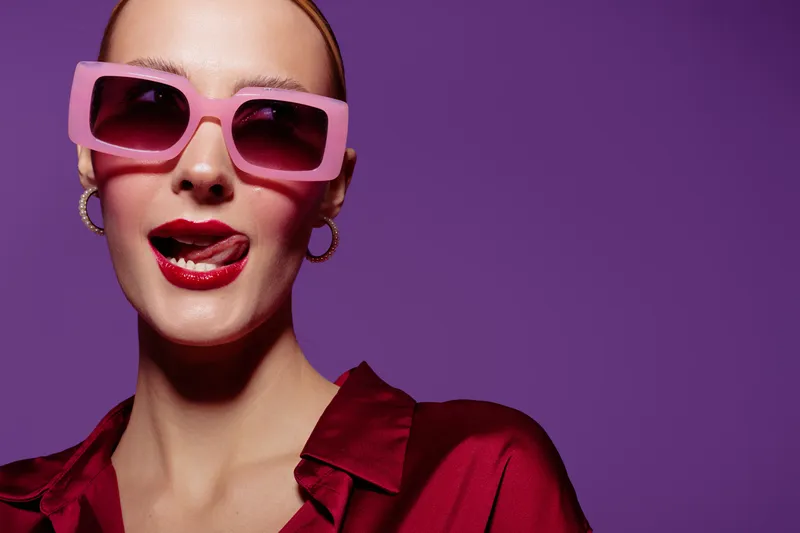 Stylish woman in pink sunglasses and red satin blouse poses with tongue out on purple background. Concept for fashion, beauty, attitude, and modern lifestyle portrait.