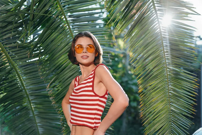 A stylish woman wearing a striped crop top and sunglasses stands amid lush palm trees, enjoying sunny outdoor vibes in a tropical setting.